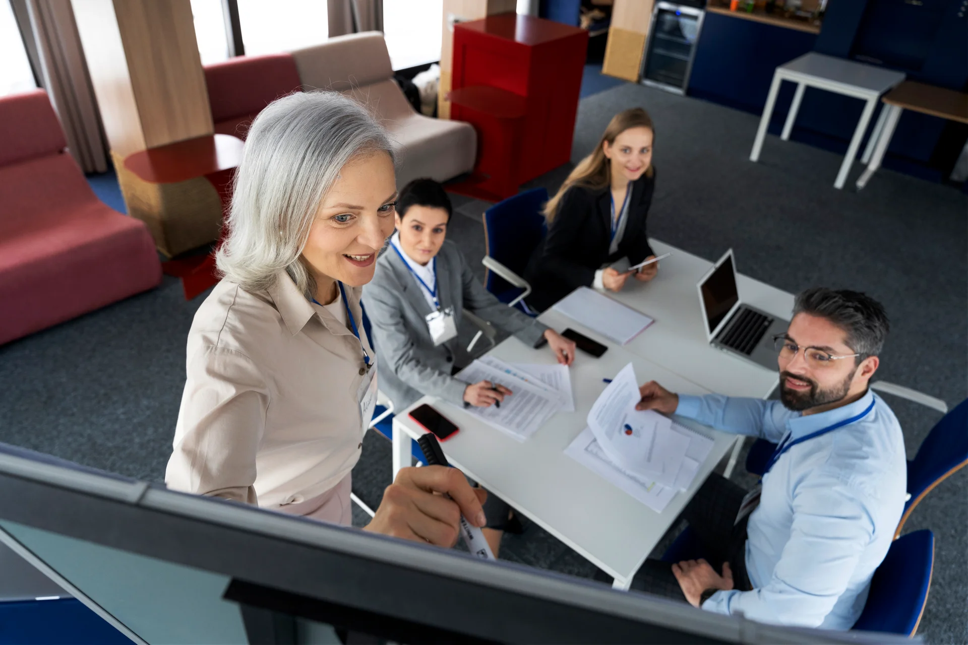 Senior female executive assistant writing on whiteboard while colleagues smile and take notes, collaborating on creating helpful resources and guides for executive assistants